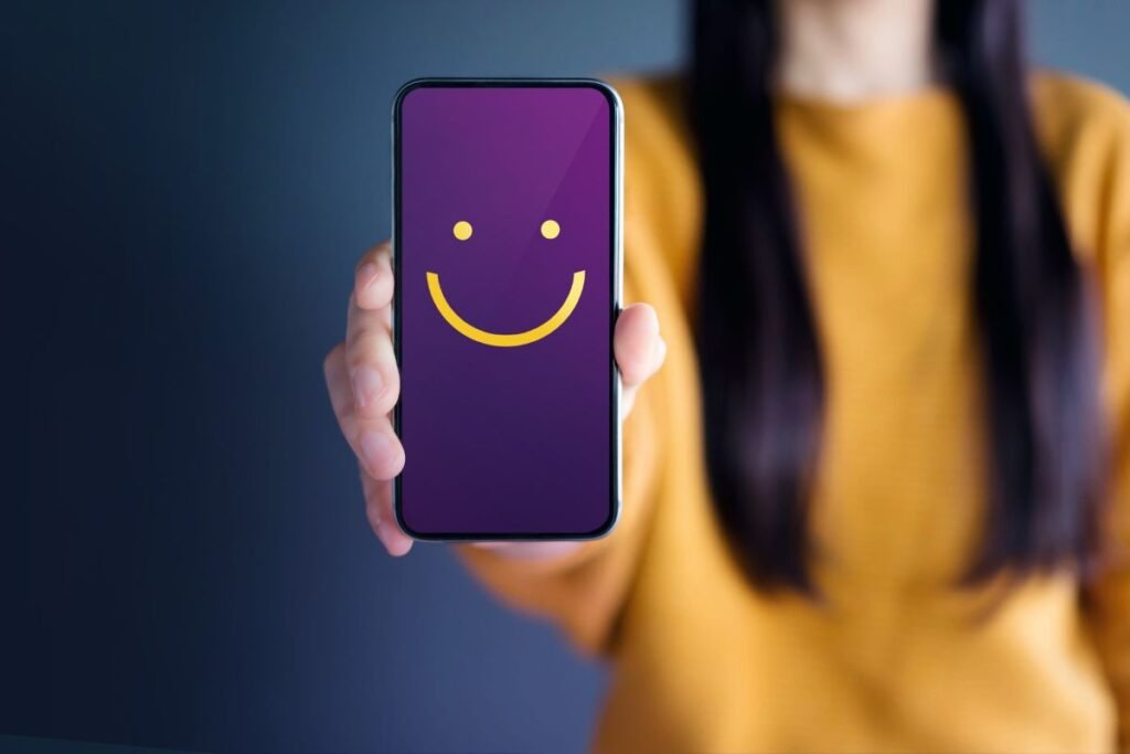 a photo if a woman in a yellow jumper holding a phone with a purple screen displaying a smiley face